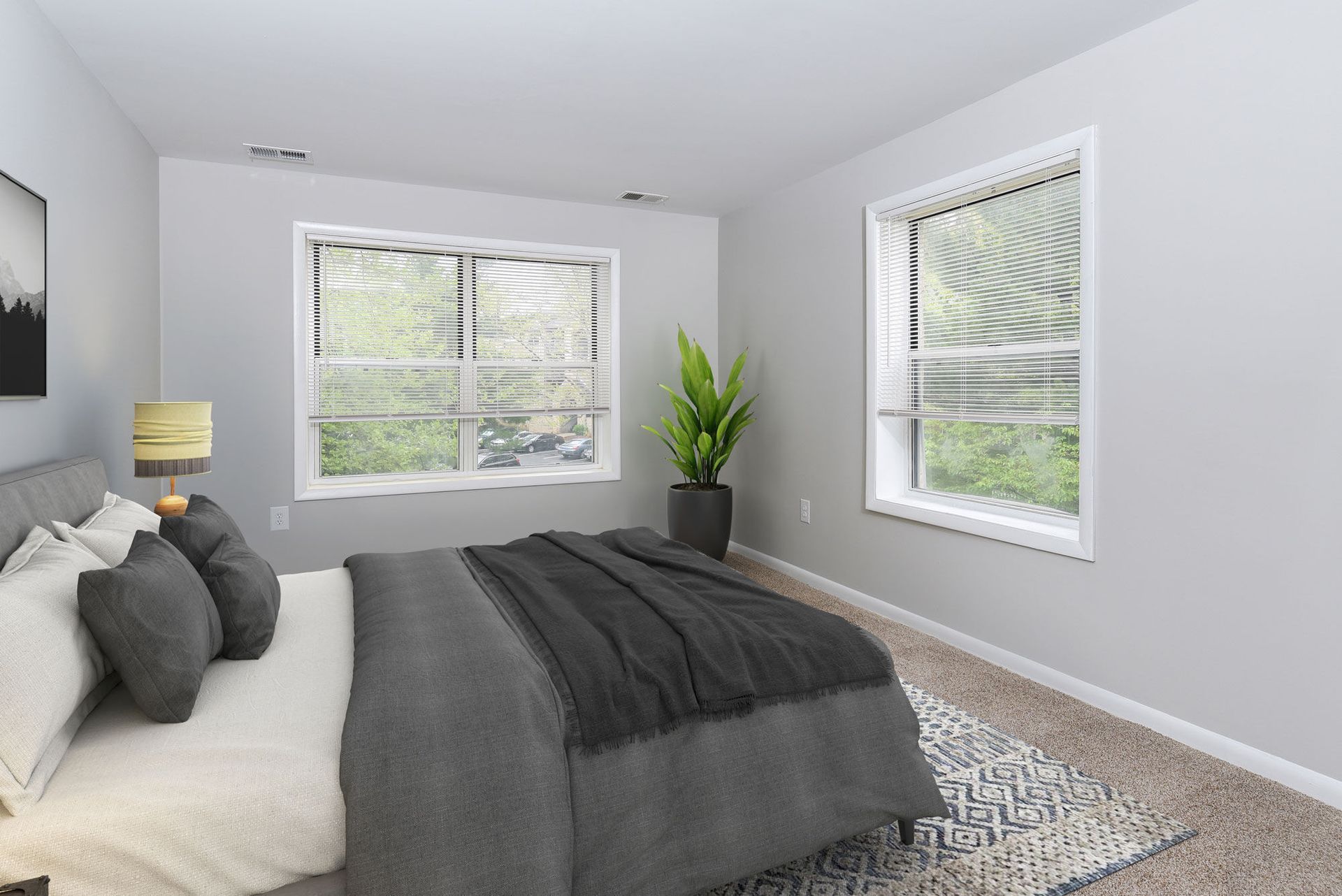 Furnished bedroom in an apartment with two large windows, gray bedding, and a potted plant.