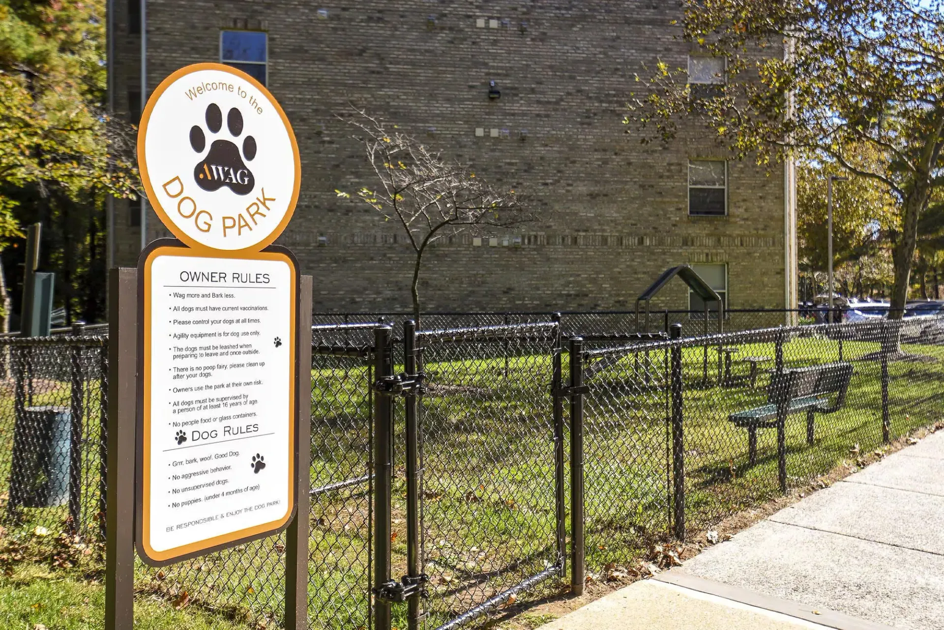 Dog park sign with paw print and rules beside a chain-link fence at an apartment complex.