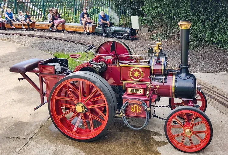 A red and black tractor is parked next to a small train.