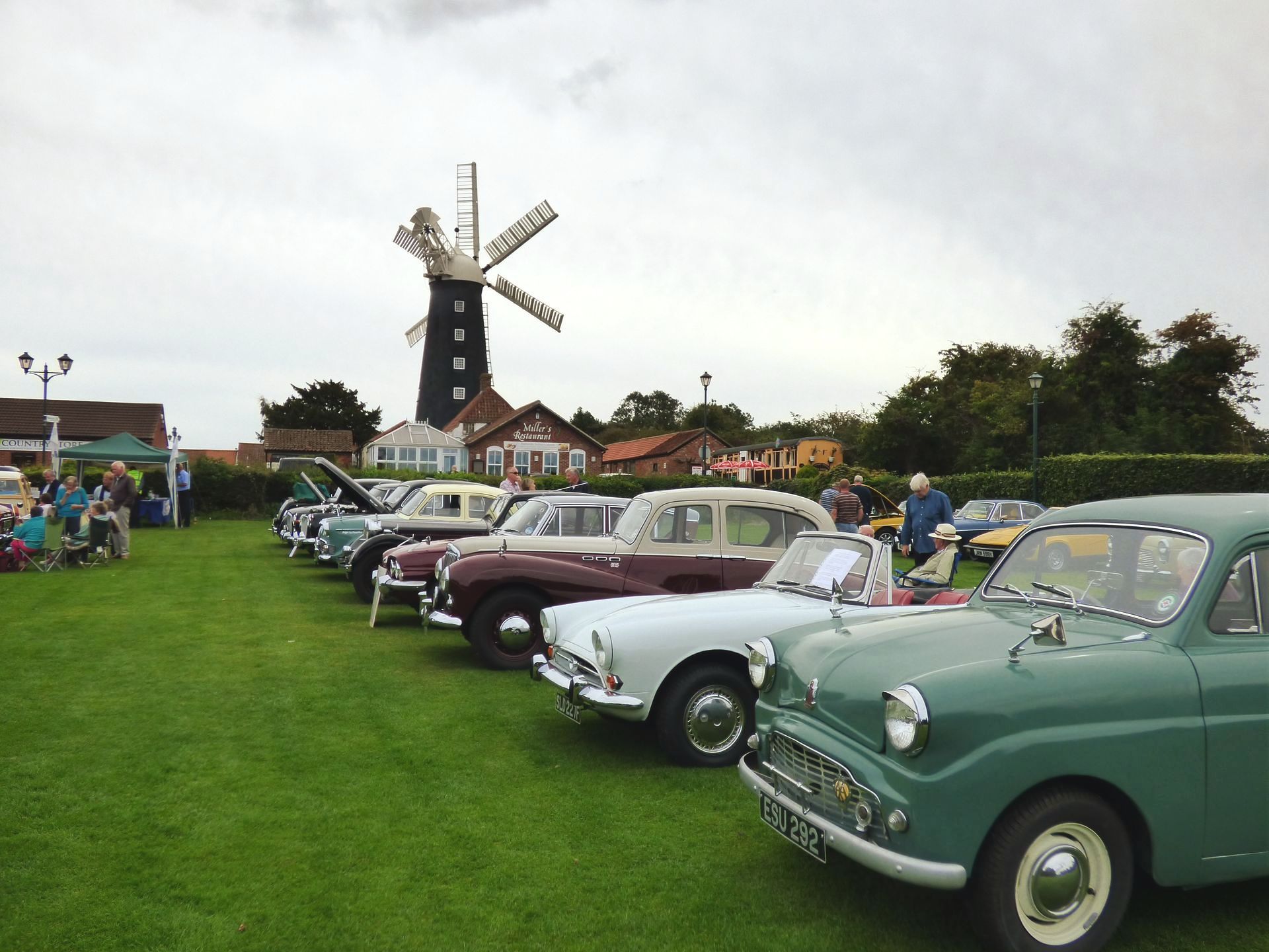 A row of old cars are parked in a grassy field with a windmill in the background