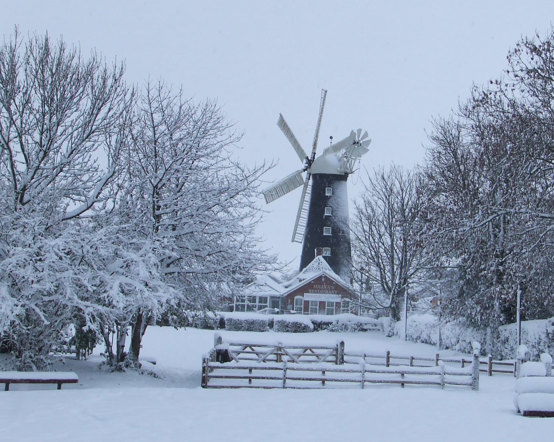 A windmill is surrounded by snow covered trees and a fence