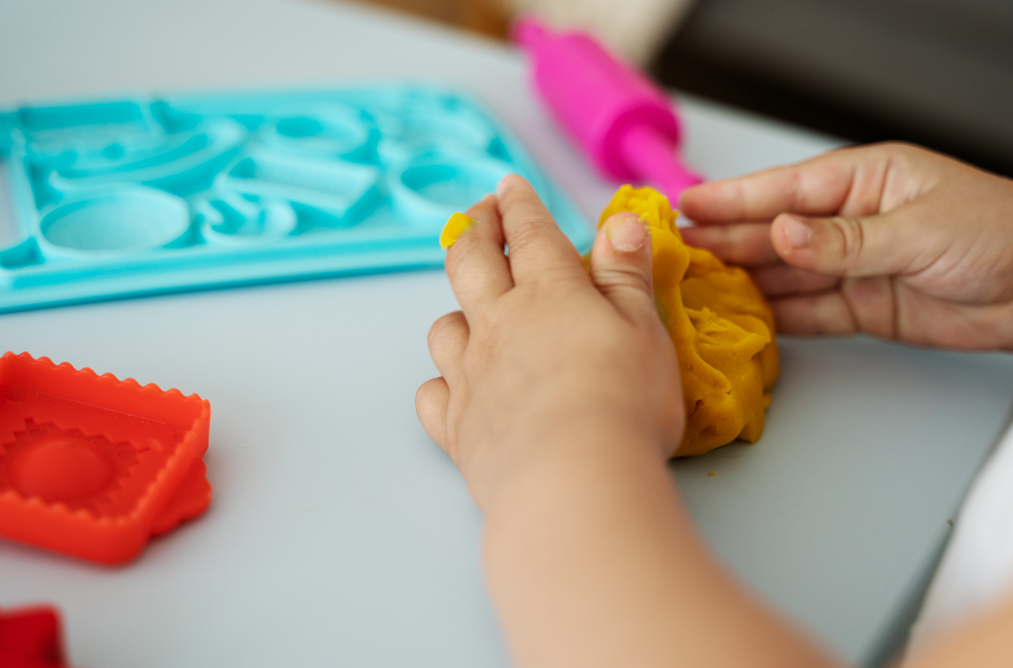 Mujer mayor sonriente con gafas en una clase de arte, rodeada de otras personas, con obras de arte coloridas en el fondo.
