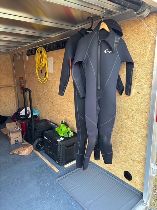 Two black wetsuits hang from a rail inside a storage trailer with a yellow circular life ring and cart in the background.
