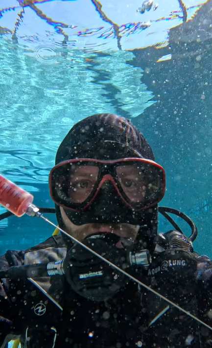 A person in scuba gear, including a mask and hood, takes a selfie underwater with bubbles floating around them.
