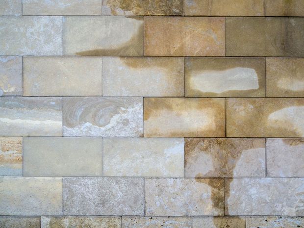 A wall of rectangular, textured stone blocks in varied shades of tan, beige, and gray with some damp, darker patches.