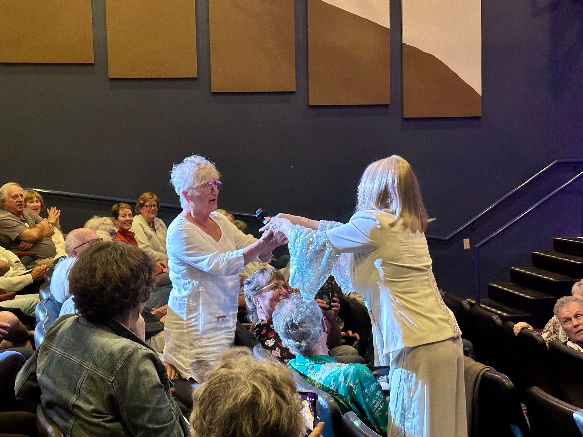 Two women in white clothing on stage, gesturing to an audience in a theater.