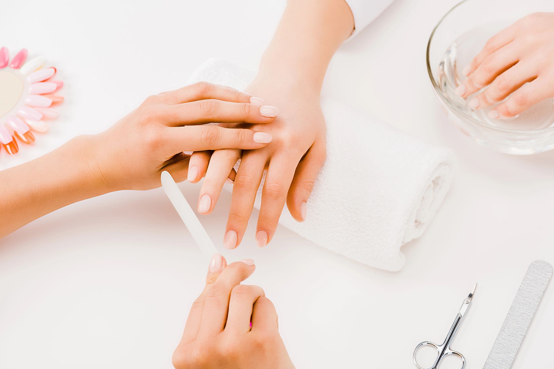 Manicure che limatura le unghie della cliente su un tavolo bianco con attrezzi, ciotola per l'acqua e asciugamano.
