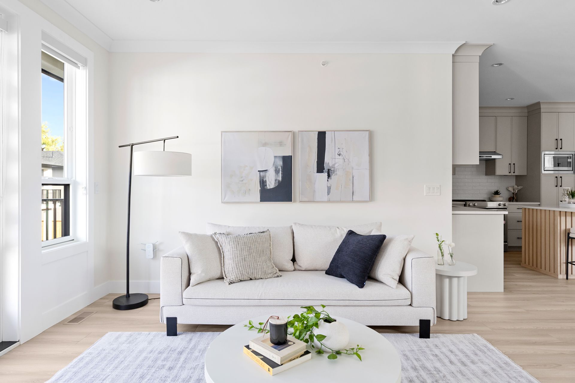 Bright modern living room with white sofa, floor lamp, abstract wall art, and light wood kitchen in background