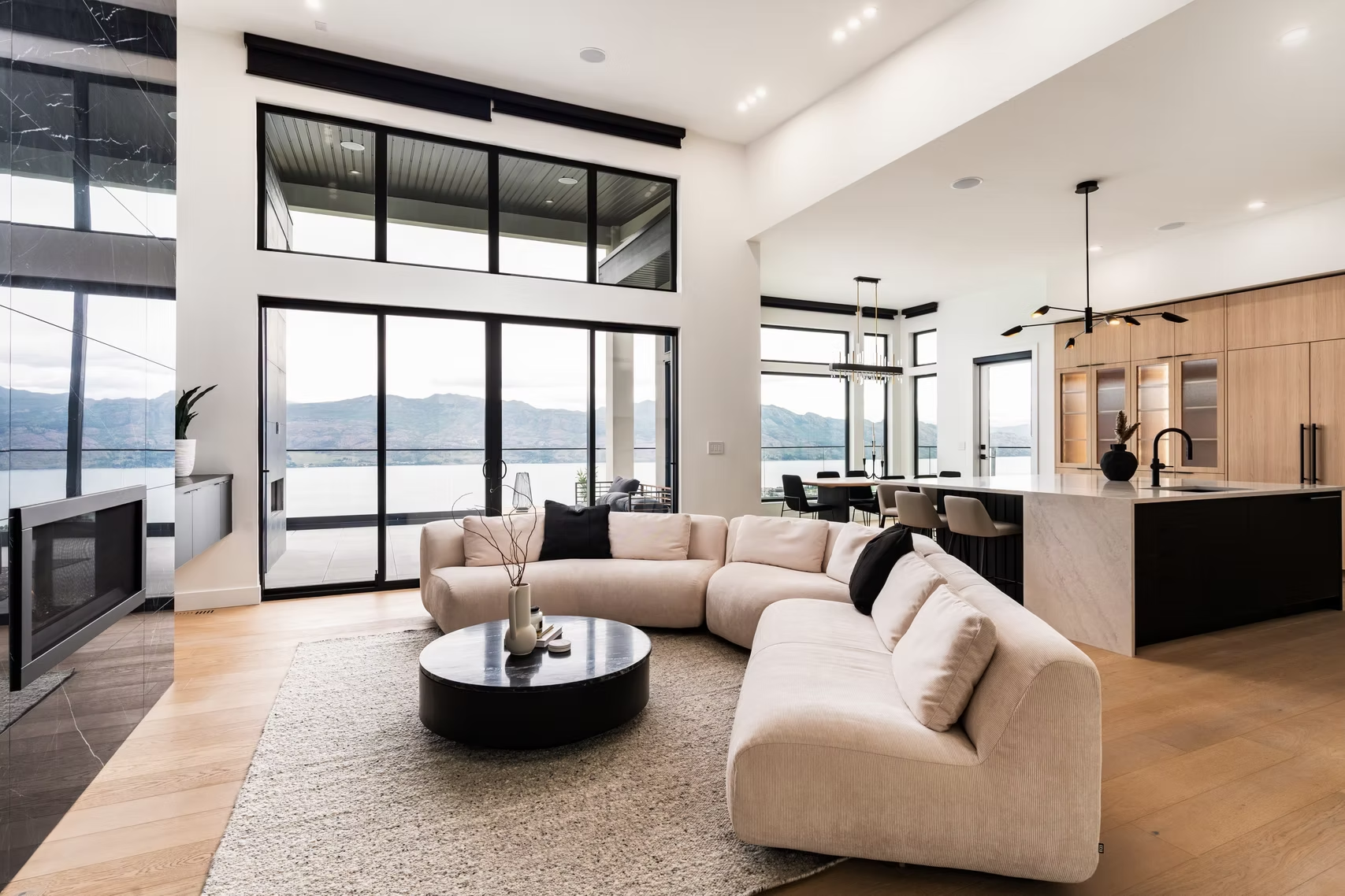 Bright modern living room with curved beige sofa, round coffee table, kitchen island, and large windows with mountain view
