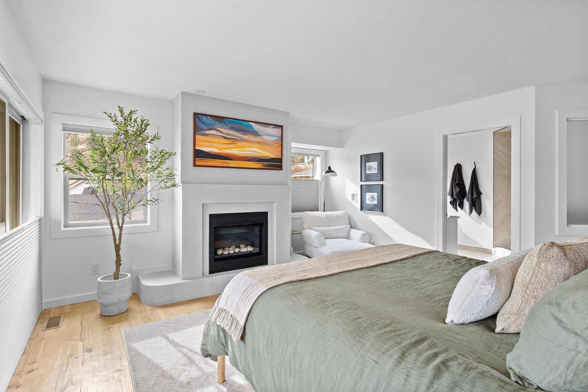 Bright bedroom with a bed, fireplace, windows, and a potted tree near a framed painting.