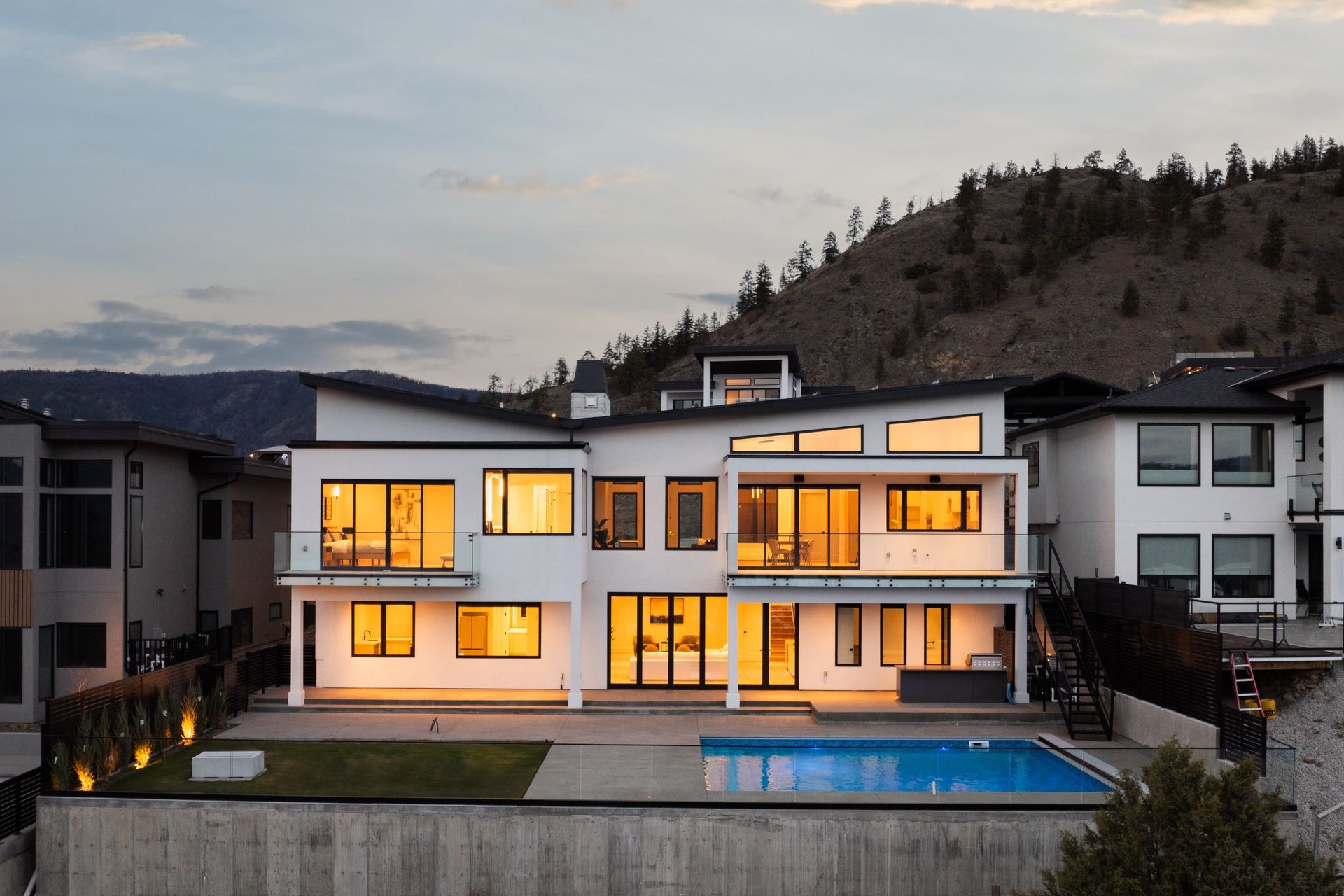Modern white hillside home at dusk with lit windows and a backyard pool