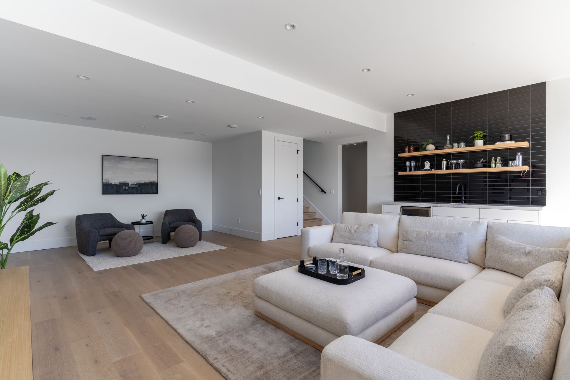 Modern living room with beige sectional sofa, lounge chairs, and a dark built-in shelf against a brick wall.