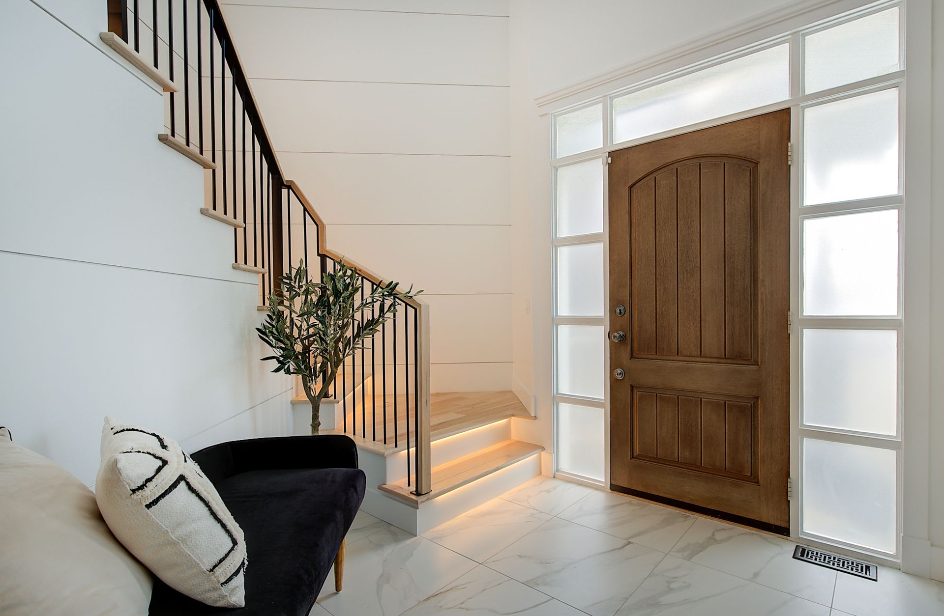 Bright entryway with wooden front door, staircase, bench, and potted plant in a white interior.