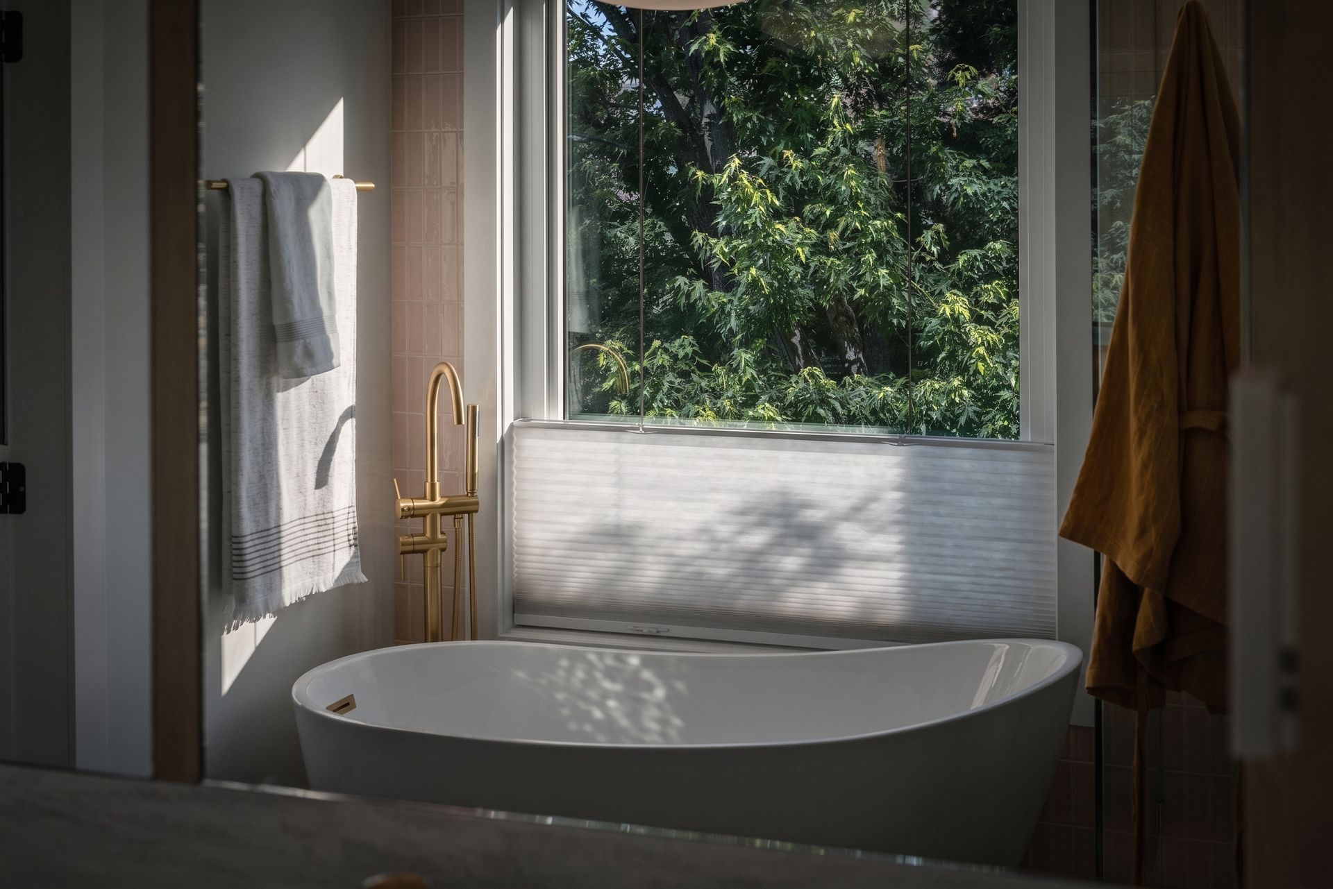 Sunlit bathroom with a freestanding tub by a window, framed by curtains and greenery outside
