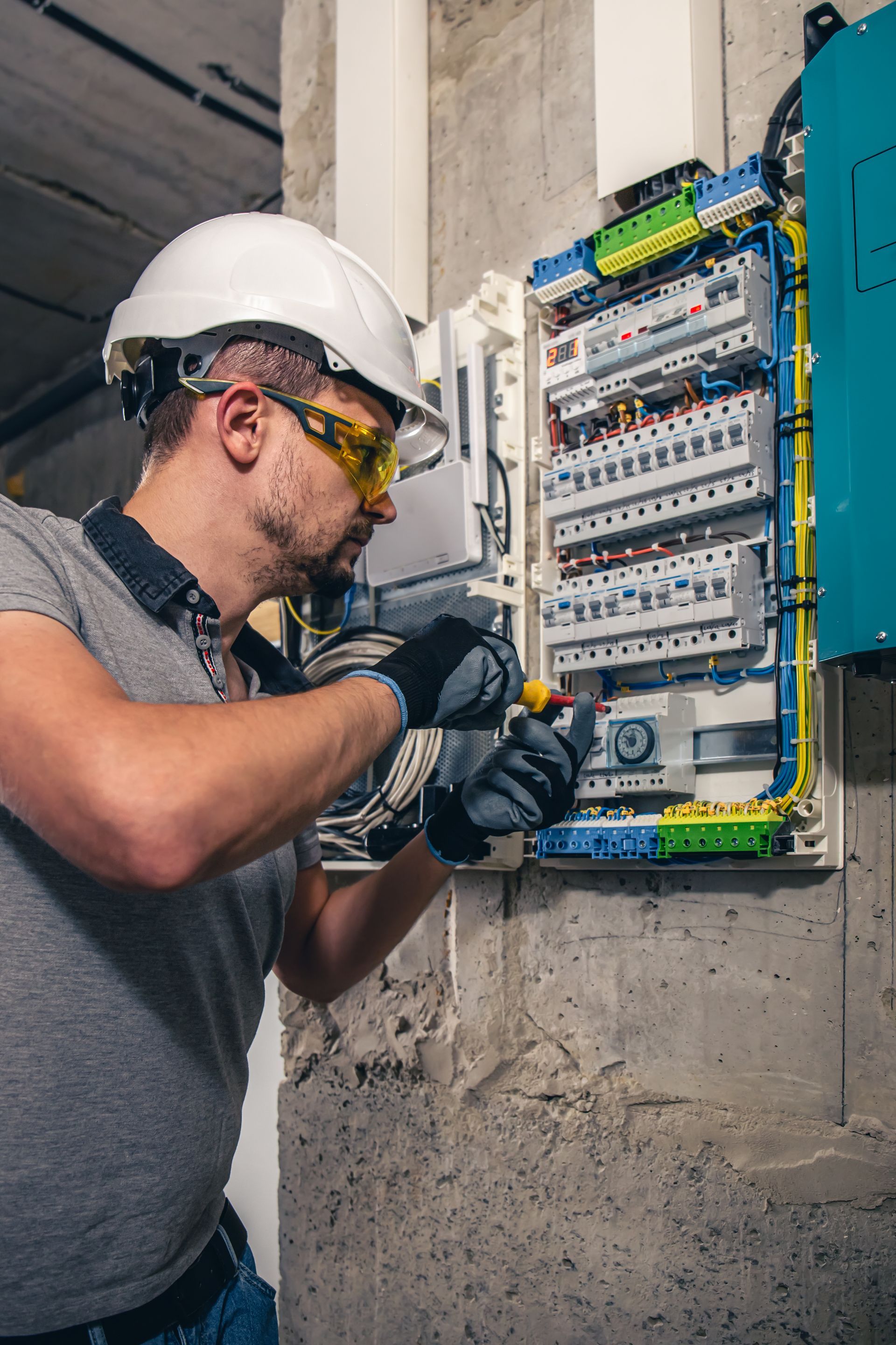 Electrician wiring a circuit breaker panel indoors, wearing a hard hat and safety glasses.