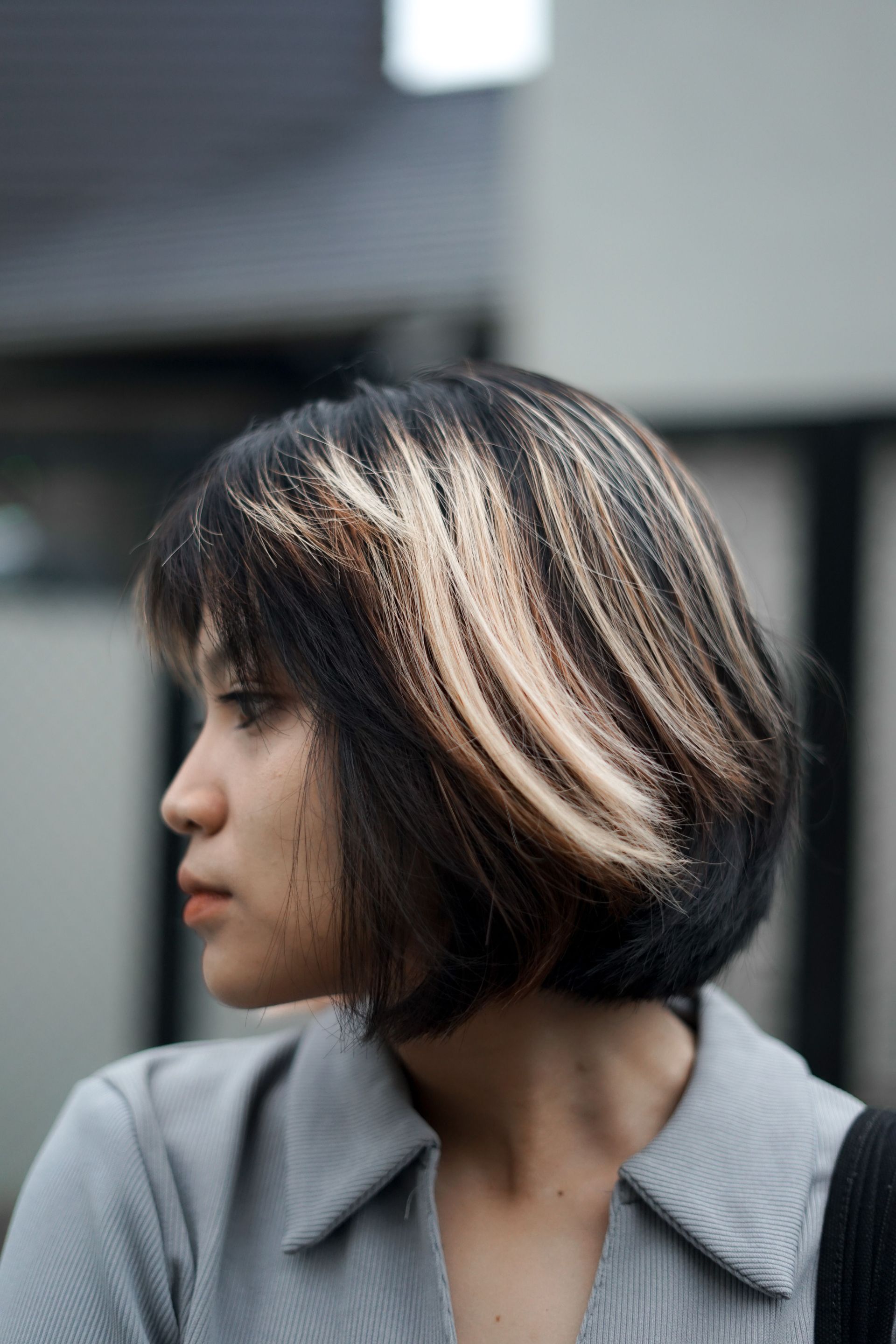 Profile view of a person with a short brown bob haircut, wearing a gray collared shirt, against a blurred outdoor background