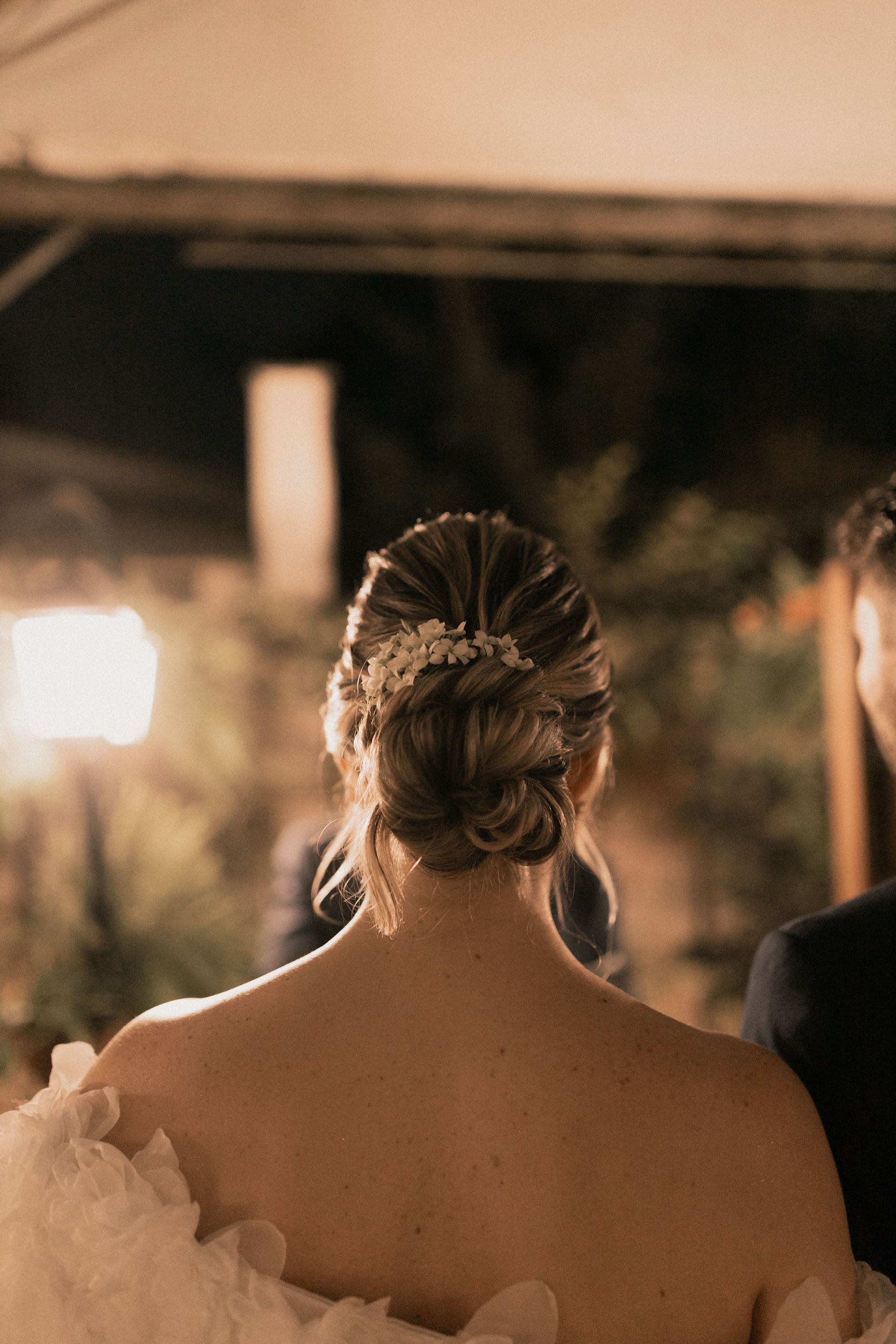 Bride seen from behind in a white dress and updo, standing outdoors at night with warm lights in the background