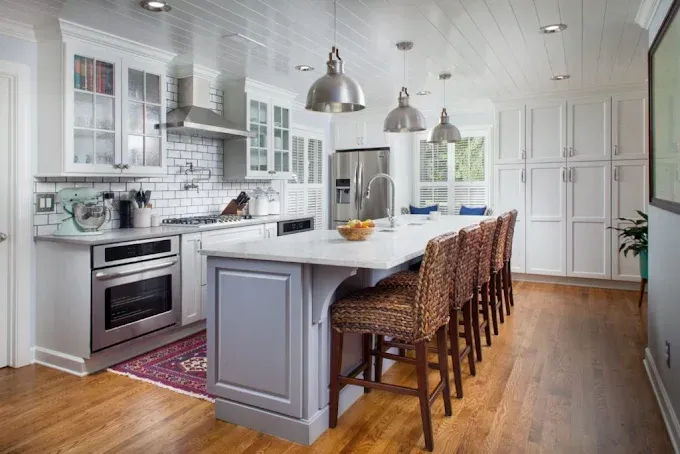 Spacious white kitchen with island, stainless steel appliances, and wooden floor. Wicker barstools line the island.