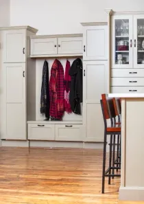 Light-colored wooden built-in entryway storage with coats hanging and a cabinet; visible kitchen counter and stools.