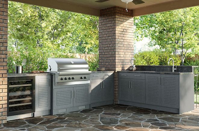 Outdoor kitchen with grill, cabinets, sink, and wine cooler. Stone patio and lush greenery.