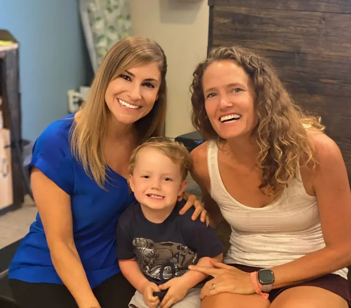 Two smiling people pose with a young child between them in an indoor setting with wood-paneled walls.