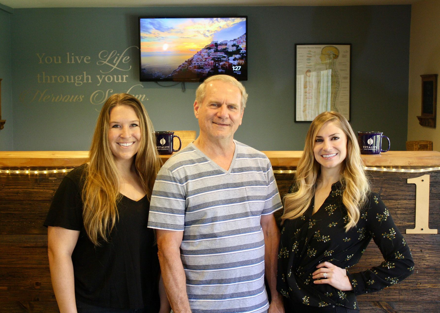 Three people smiling in front of a wood-paneled reception desk with a television and wall art in the background.