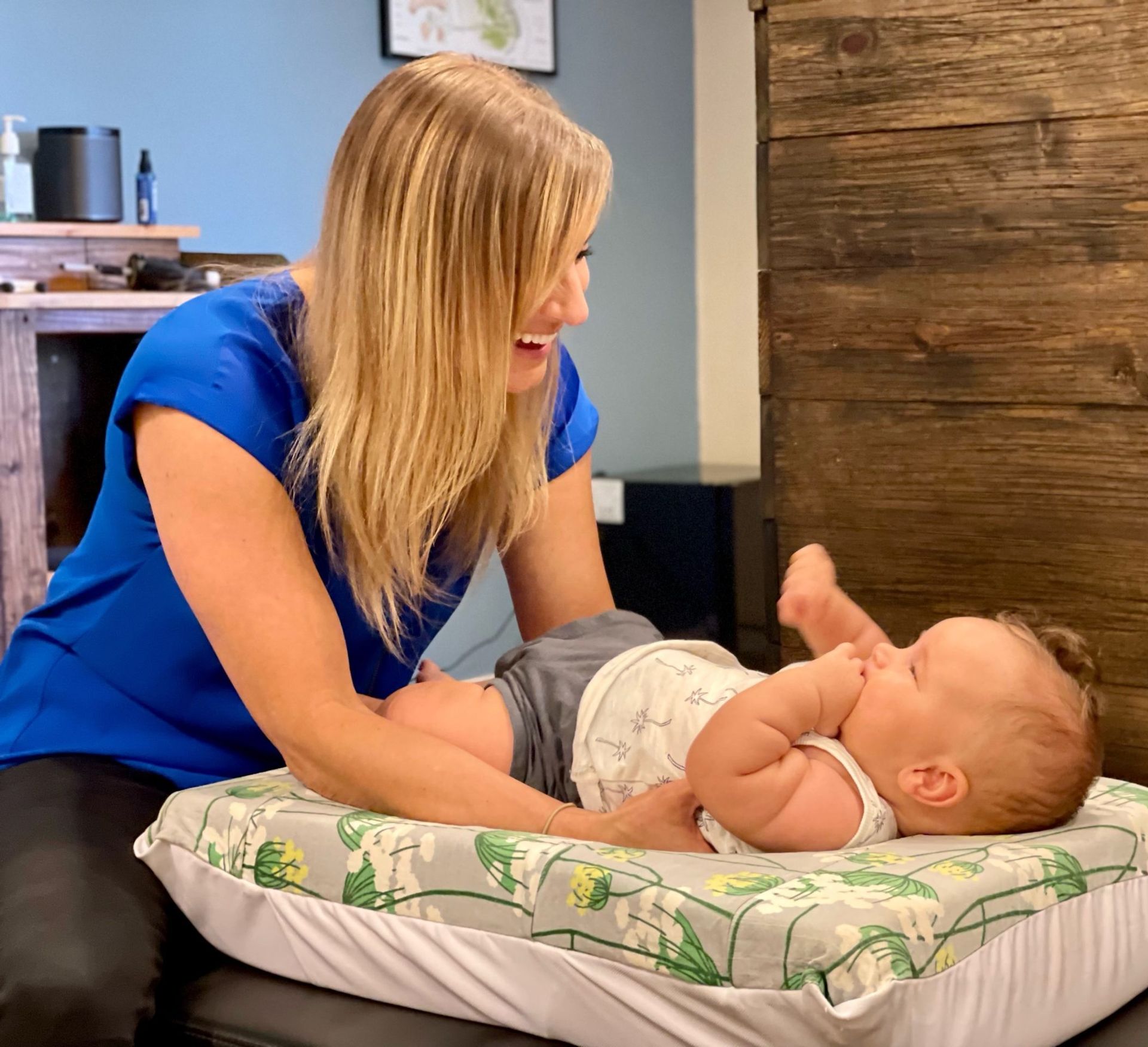 A person in a blue shirt smiles while interacting with an infant lying on a floral-patterned cushion.