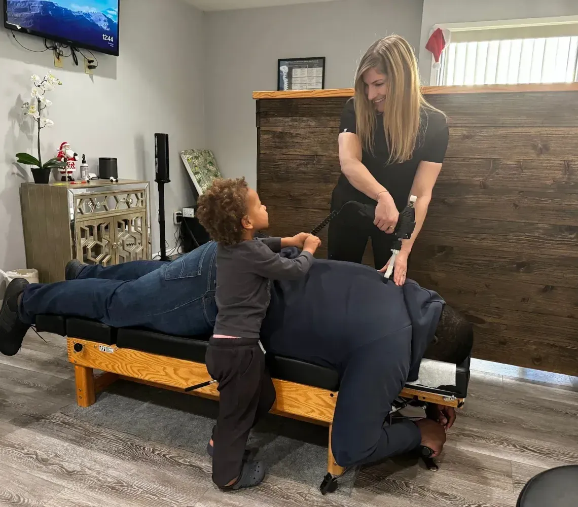 A practitioner uses an adjustment tool on a person's back while a child observes from beside the treatment table.