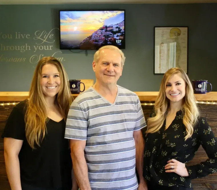Three smiling people stand behind a wood-paneled reception desk with an inspirational quote on the wall behind them.