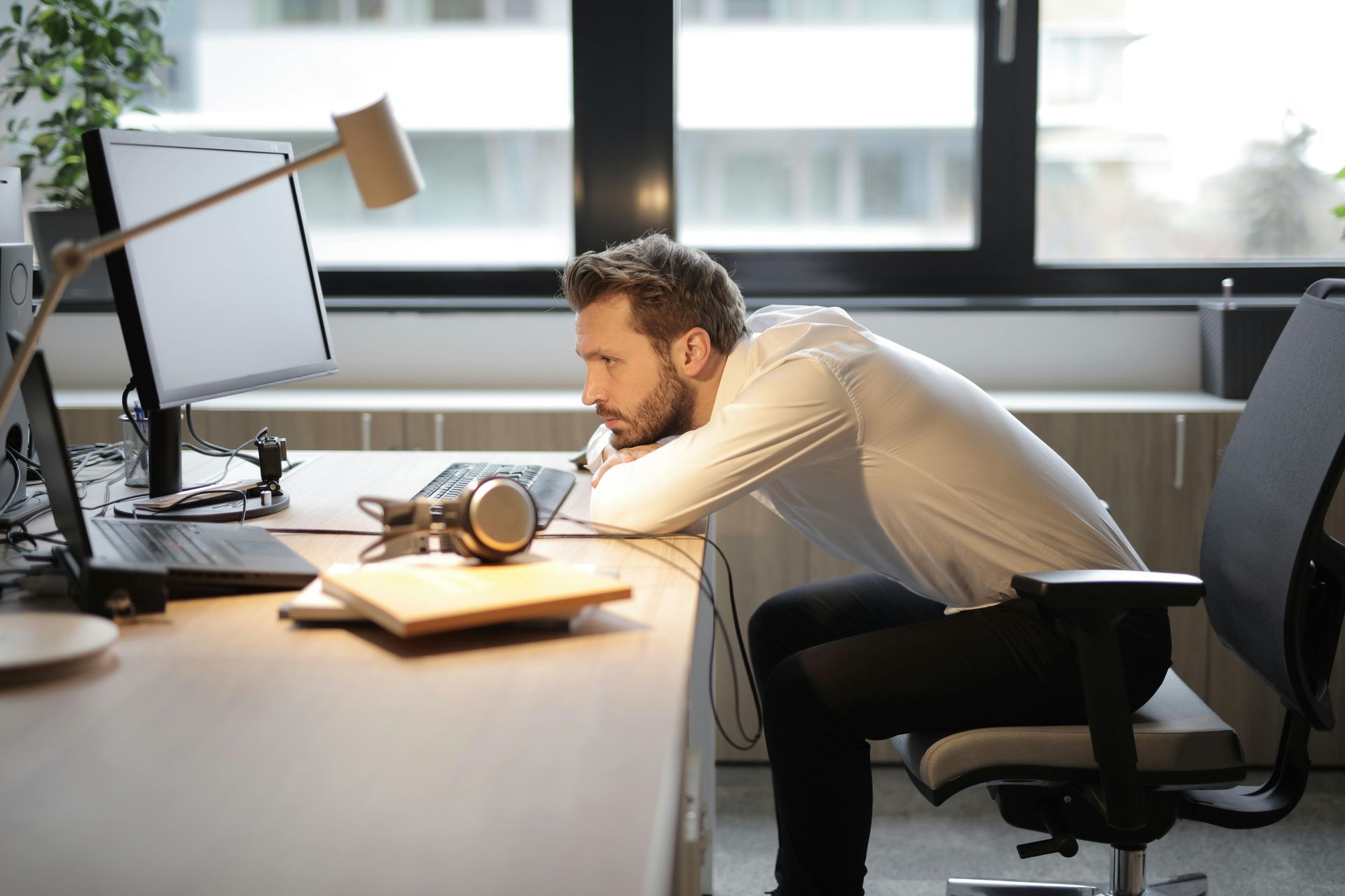 A person in a white shirt leans tiredly on a desk, staring intently at a computer screen in an office setting.
