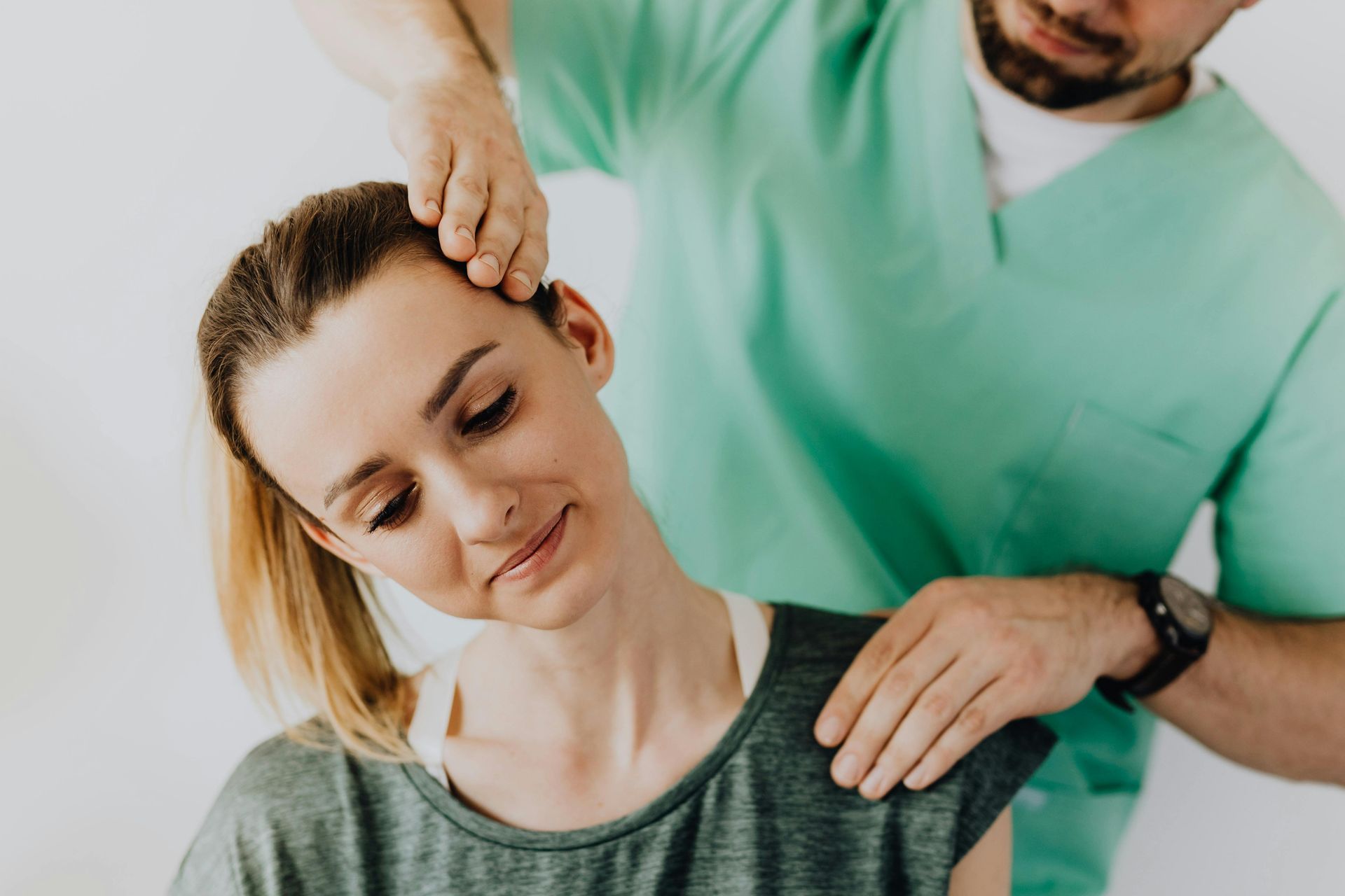 A healthcare professional in green scrubs performing a gentle neck stretch on a patient against a plain white background.