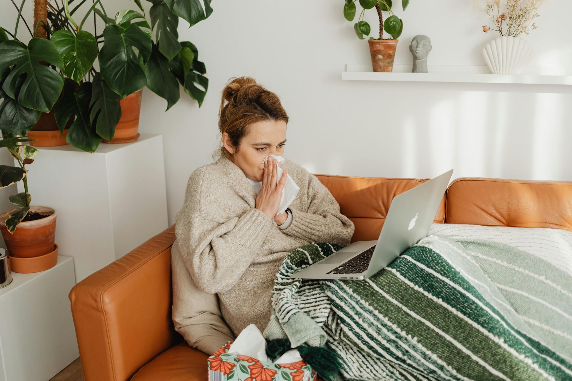A person sits on a brown sofa, wrapped in a blanket, looking at a laptop while holding a tissue to their nose.