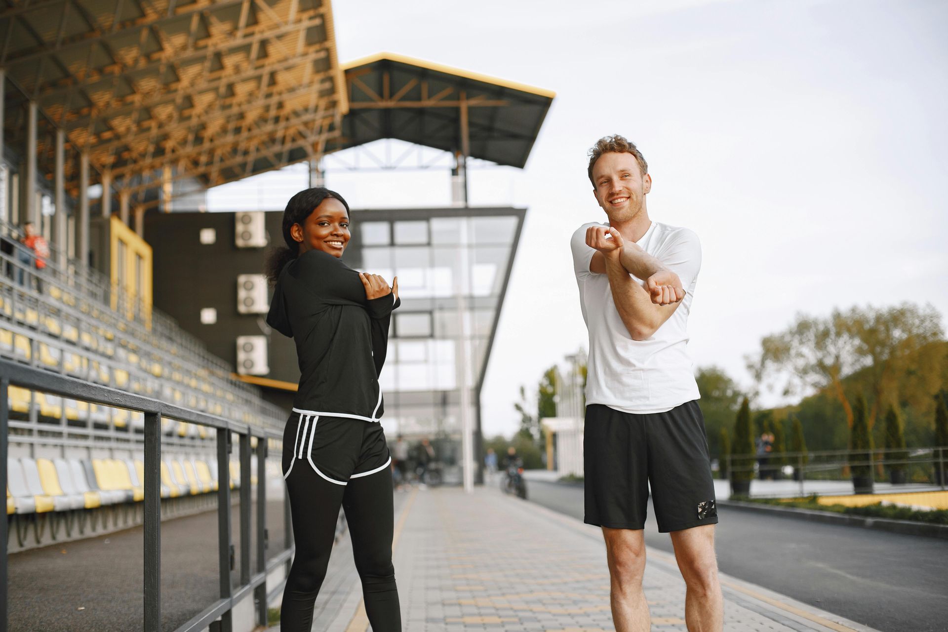 Two people in athletic wear stretching their arms on a walkway outside a stadium.