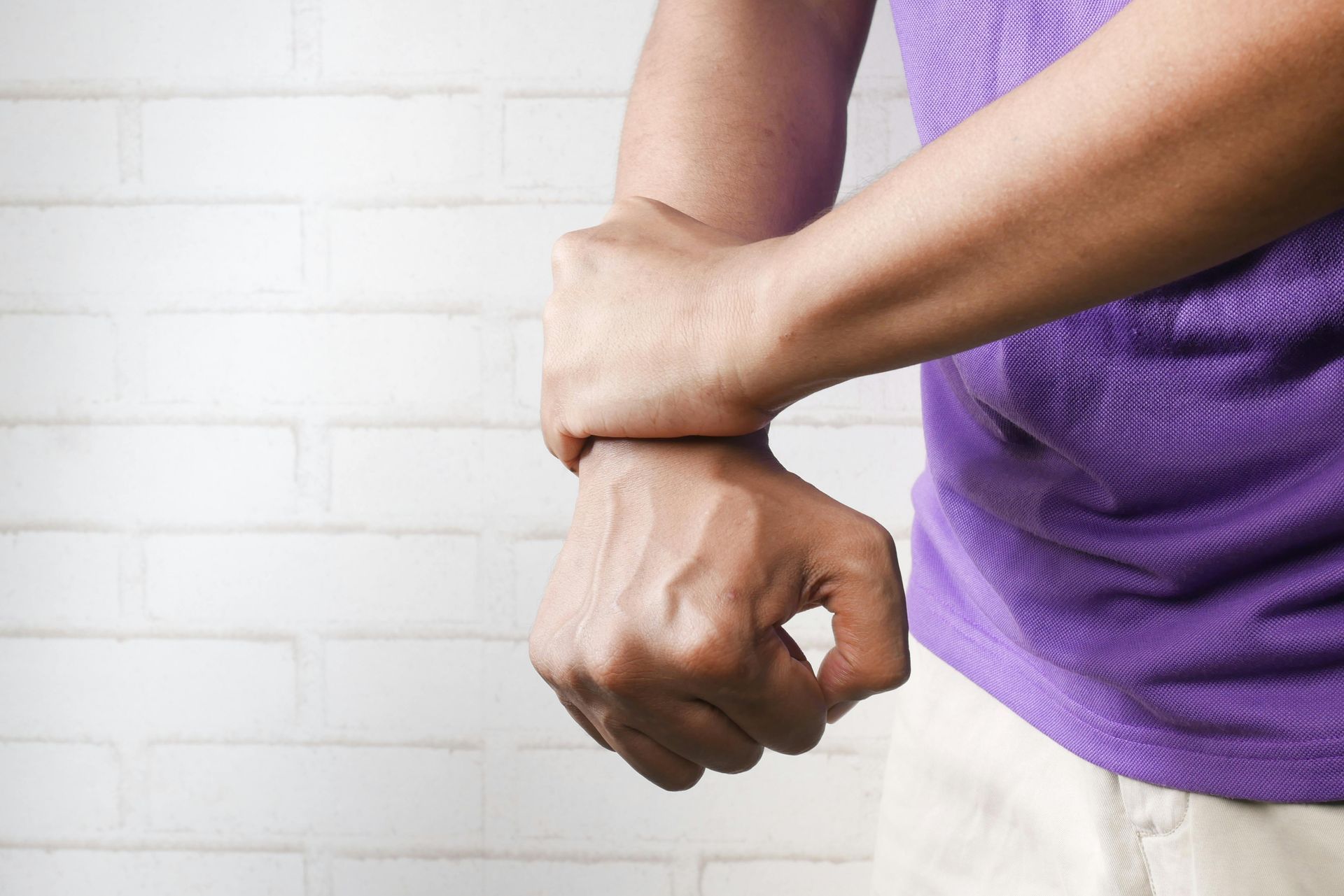 A person in a purple shirt grips their own wrist with their other hand, suggesting pain or discomfort.