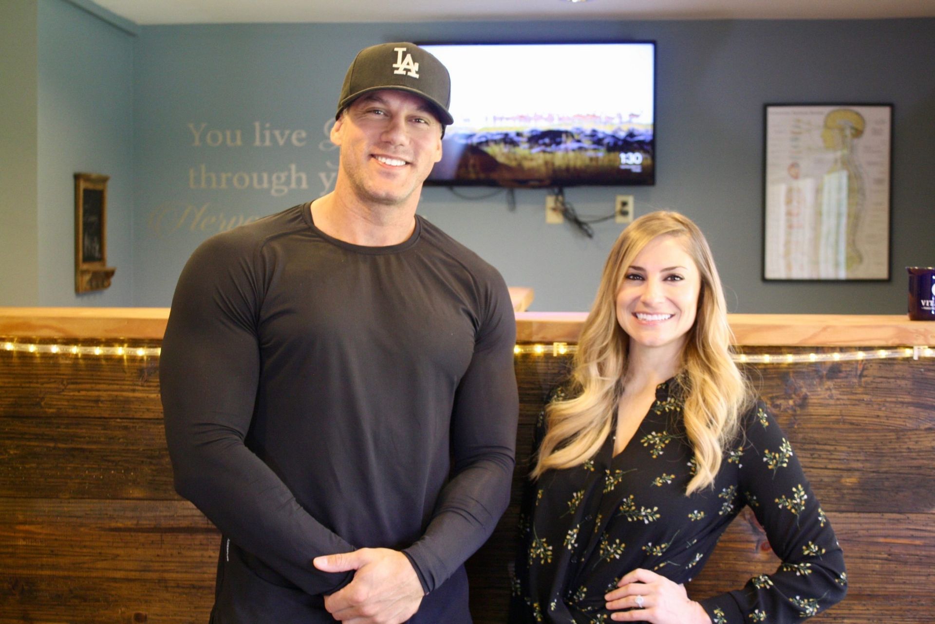 A man in a black long-sleeve shirt and cap and a woman in a floral blouse stand smiling behind a wooden service counter.