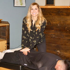 A smiling professional in a black floral blouse performs a hands-on physical therapy adjustment on a patient.