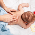 Hands gently massaging the back of a baby lying on a white surface next to a blue towel and a yellow toy.