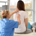 A physical therapist in blue scrubs examines the back of a person sitting on an exam table in a clinical setting.