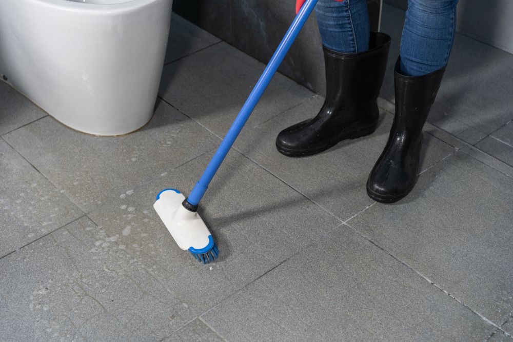 Person mopping gray tile floor with a blue-handled mop near a white fixture
