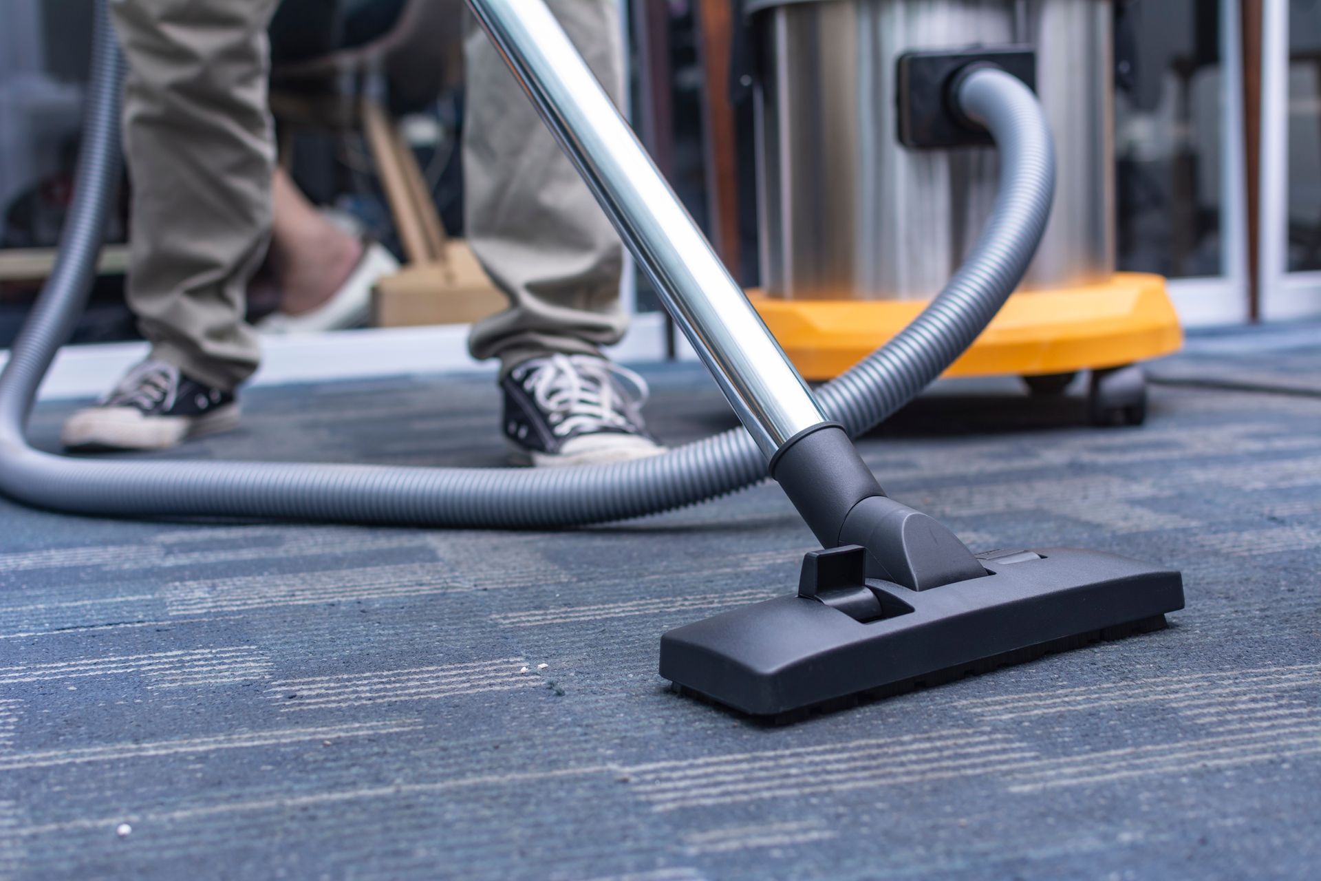 Person vacuuming a carpeted floor with a gray hose and black floor nozzle.
