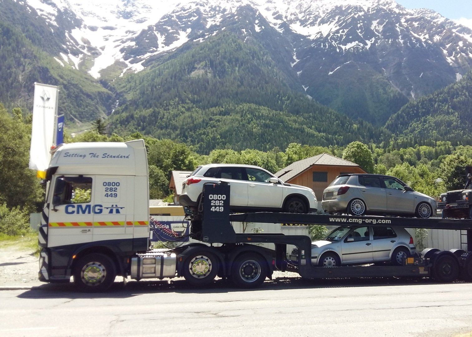 A truck carrying cars is parked on the side of the road in front of a mountain.