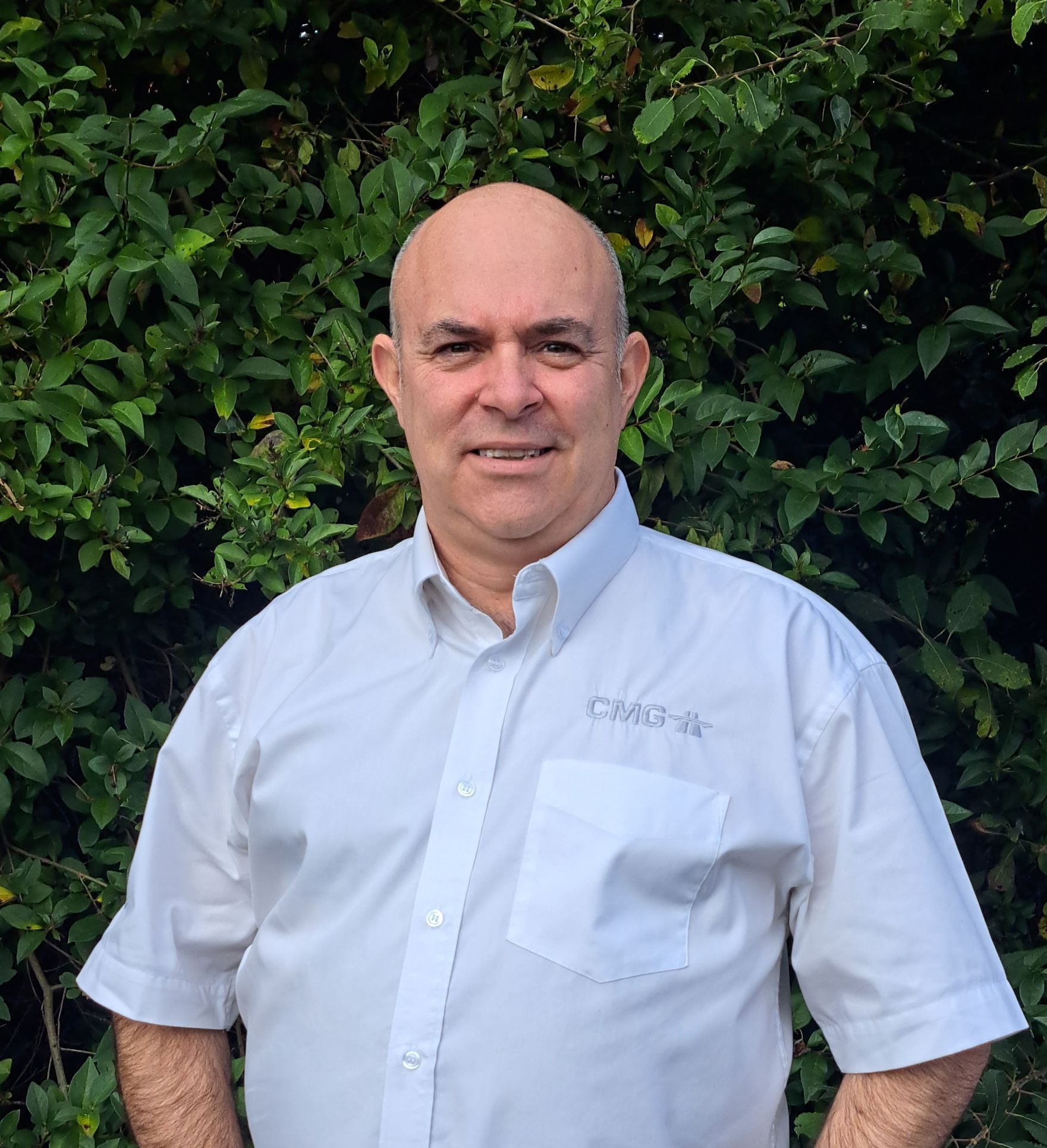A bald man in a white shirt is standing in front of a bush.
