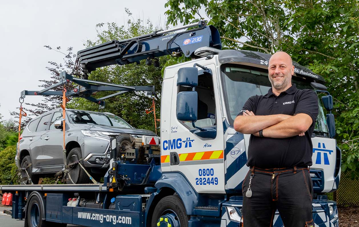 Man in front of a tow truck with a car on it. The truck is white and blue with the logo
