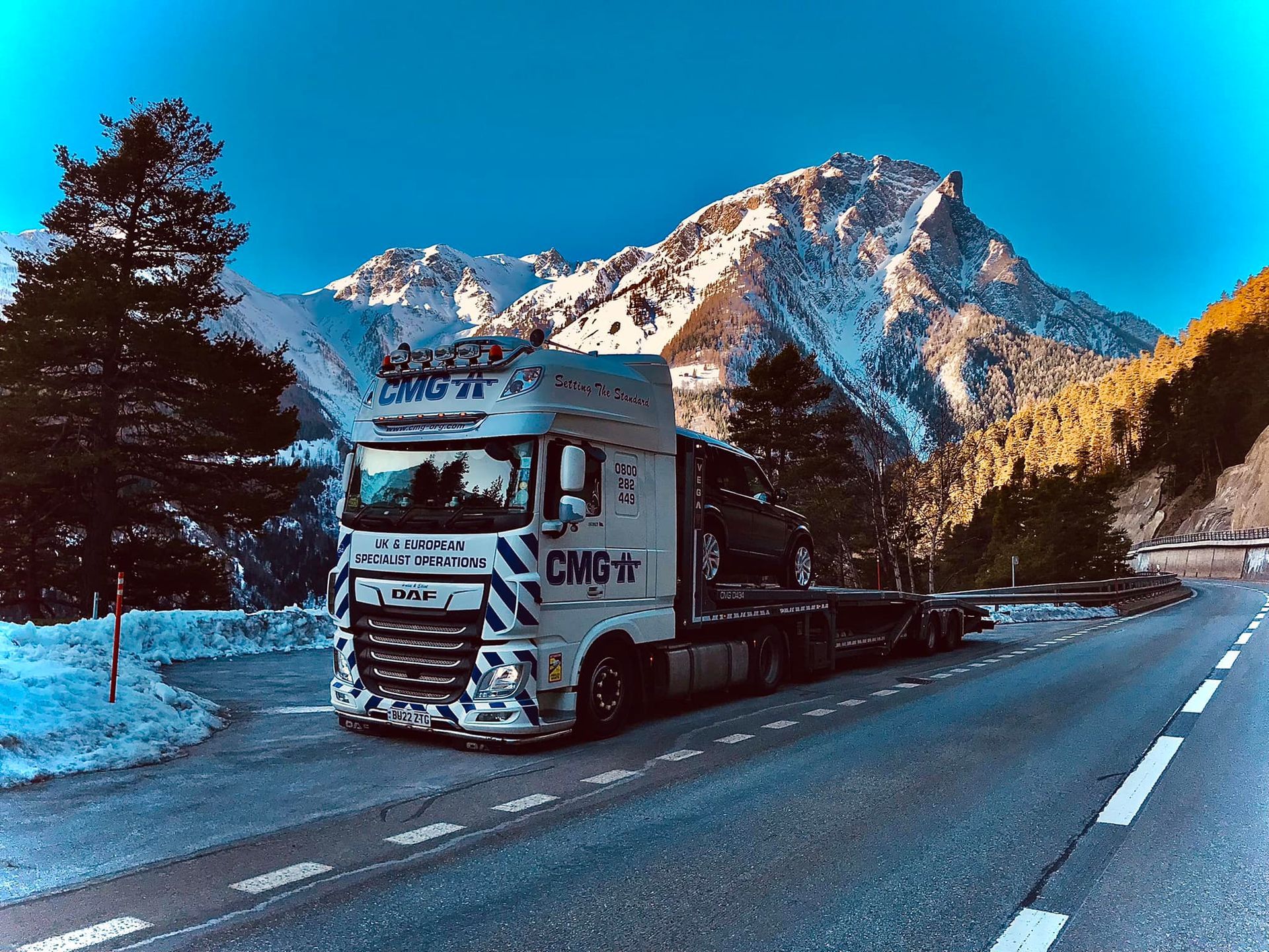 A truck is driving down a snowy road with mountains in the background.