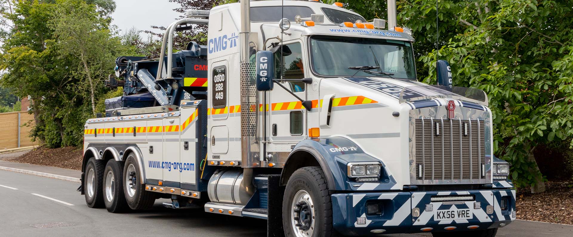 White and blue tow truck driving on a road next to some greenery.
