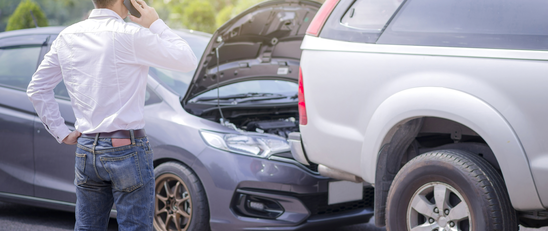 A man is standing next to a broken down car and talking on a cell phone.