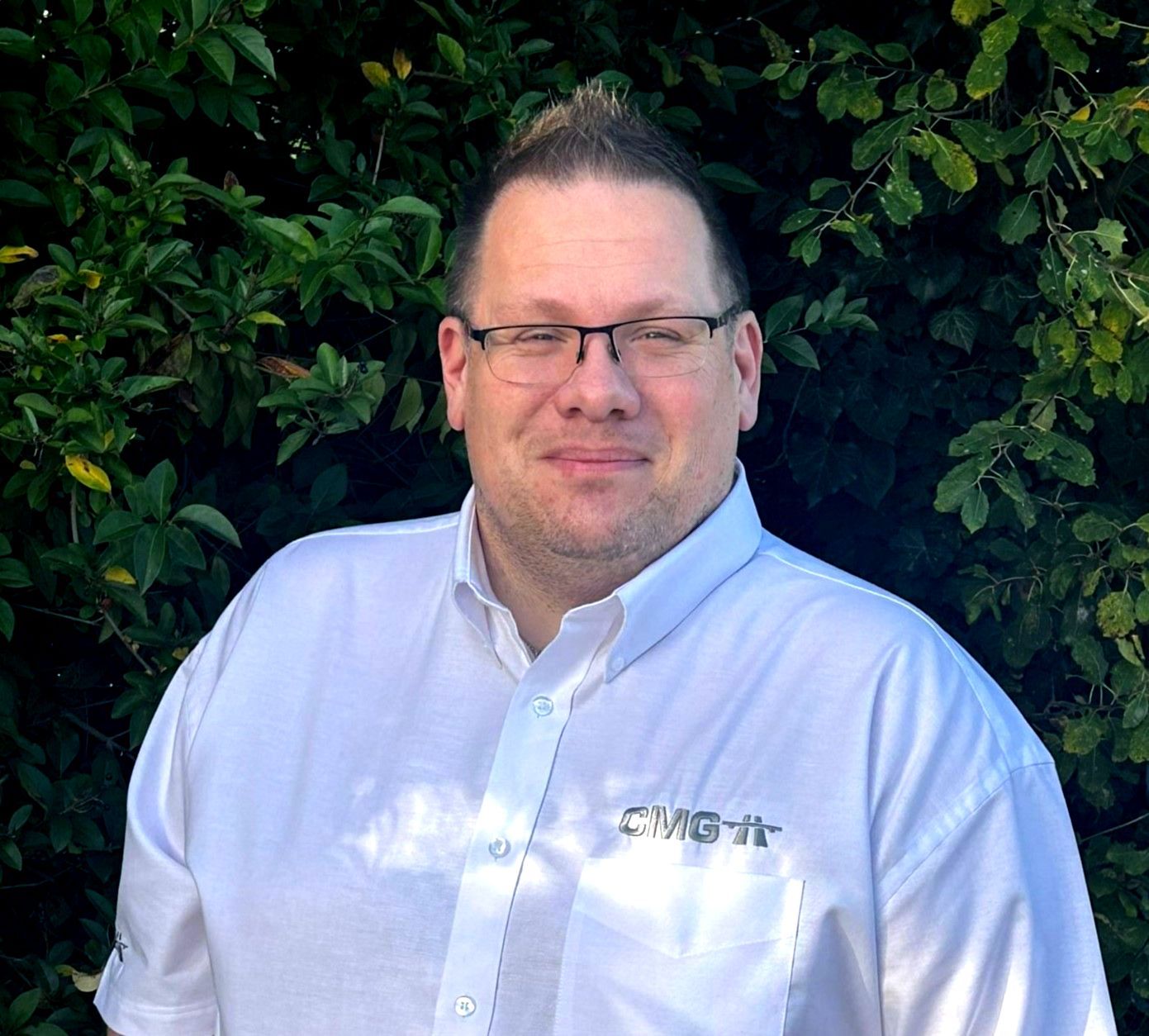 A man wearing glasses and a white shirt is standing in front of a bush.