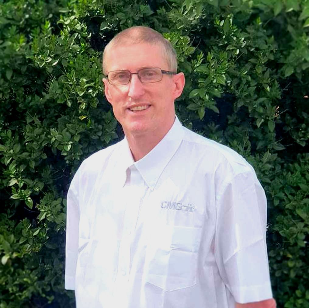 A man wearing glasses and a white shirt is standing in front of trees.