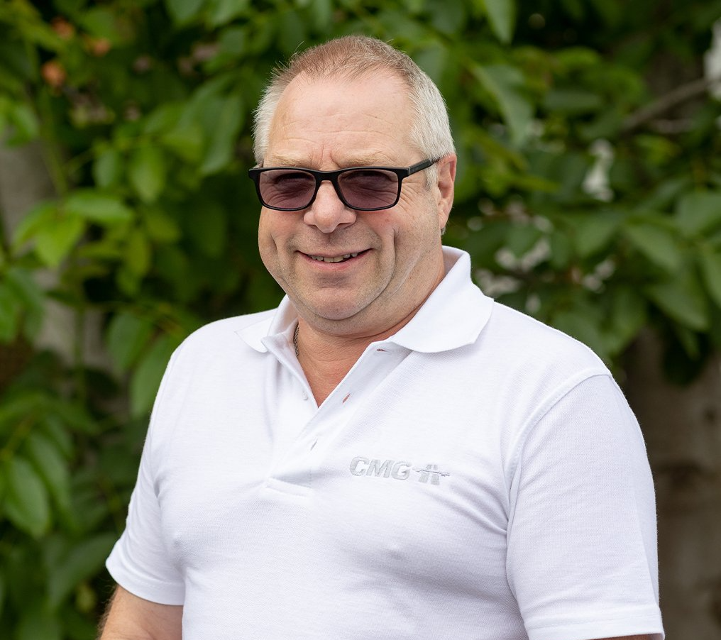 A man wearing sunglasses and a white polo shirt is standing in front of a tree.