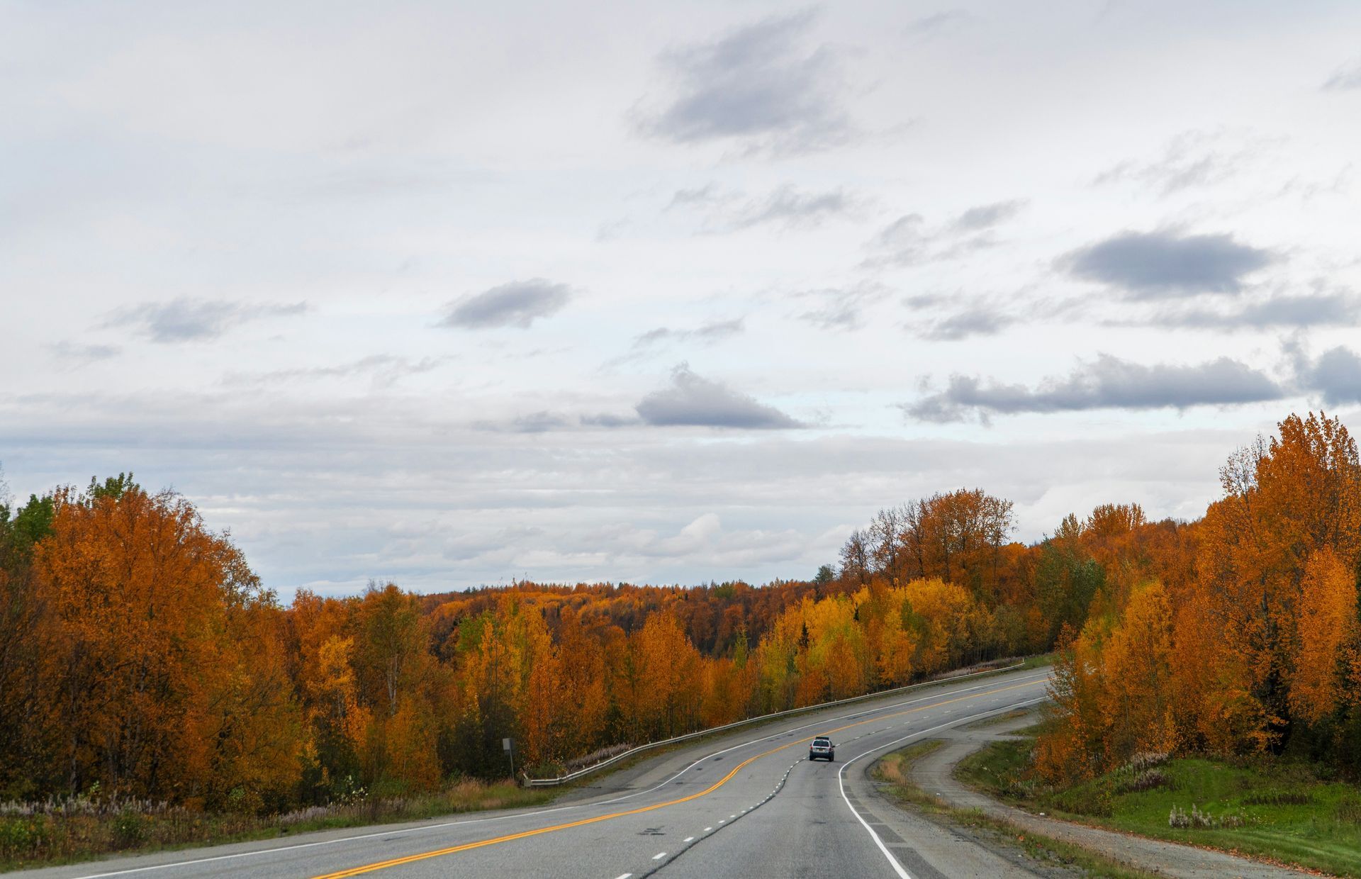 Road winding through autumn trees with a car traveling on it under a cloudy sky.